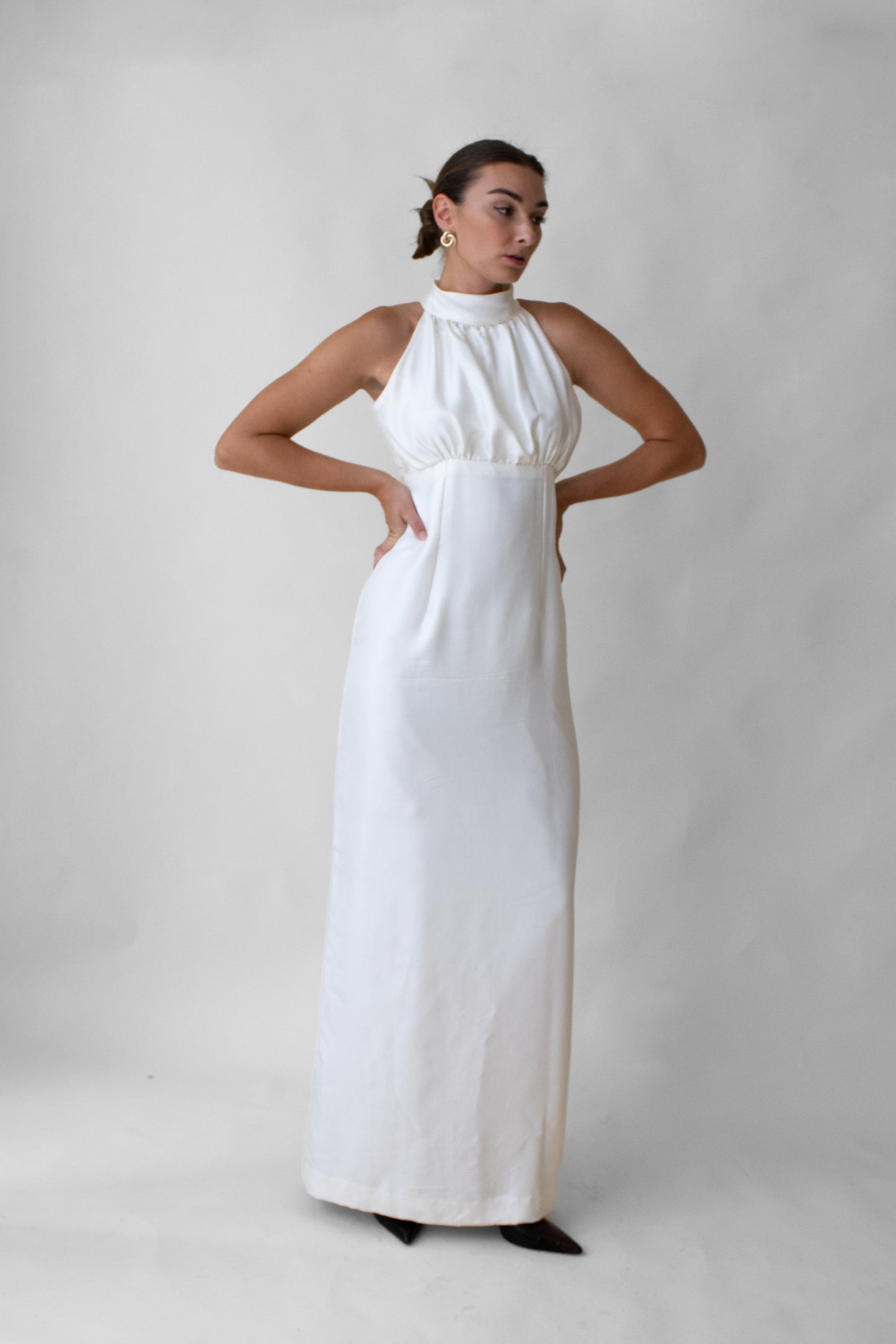 Woman wearing a white silk halter neck dress with mother of pearl button against a plain background